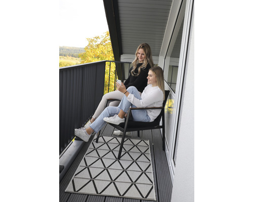 Zwei Frauen sitzen auf einem Balkon mit geometrischem Teppich und dunklem Geländer.