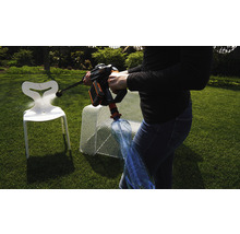 Eine Person benutzt einen Akku-Hochdruckreiniger, um einen Kunststoffstuhl im Garten zu reinigen. Eine Wasserflasche ist an den Hochdruckreiniger angeschlossen.