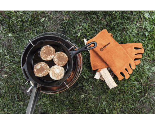 Pfannkuchen in einer gusseisernen Pfanne auf einem Kochgestell mit Petromax Logo Lederhandschuhen neben Holz.
