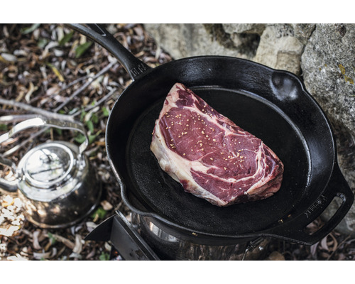 Ein rohes Steak in einer gusseisernen Bratpfanne auf einem Kocher mit einem Wasserkessel daneben
