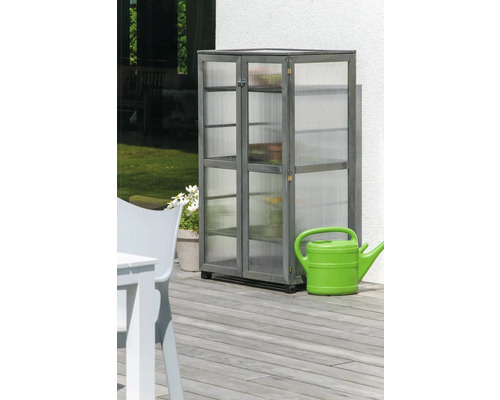 Grauer Holzschrank mit Regalen und durchsichtigen Paneelen steht auf einer Terrasse neben einer grünen Gießkanne.