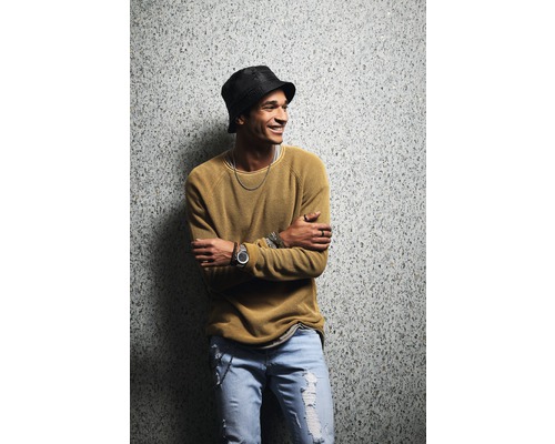 Mann mit Hut, Pullover und Jeans steht vor einer Wand