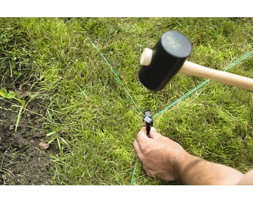 Eine Person fixiert eine grüne Richtschnur mit einem Hammer und einem Hering im Rasen.