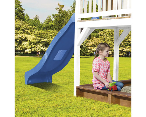 Mädchen sitzt in einem Sandkasten unter einem Spielturm mit Rutsche auf einem Rasen