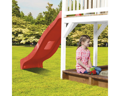 Kind im Sandkasten mit Spielzeug unter einem Spielturm mit roter Rutsche