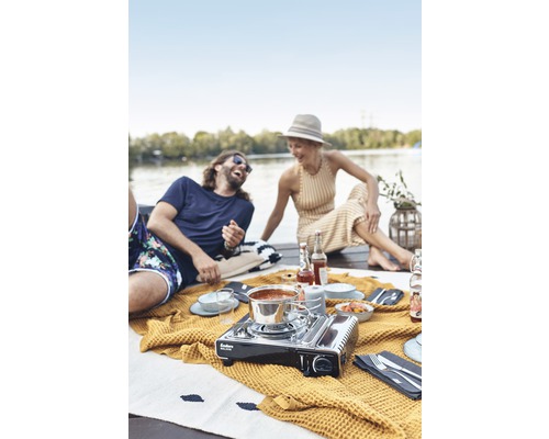 Szene mit Personen, Picknickdecke und einem tragbaren Gaskocher für den Außenbereich.