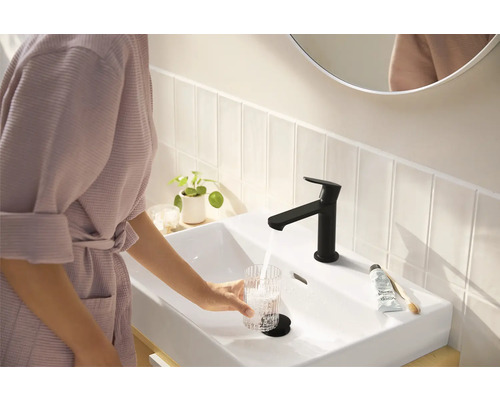 Frau füllt Glas Wasser an einem Waschbecken mit schwarzer Armatur in einem hellen Badezimmer.