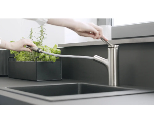 Eine Person gießt mit einer Küchenarmatur Kräuter in einem Pflanzgefäß.