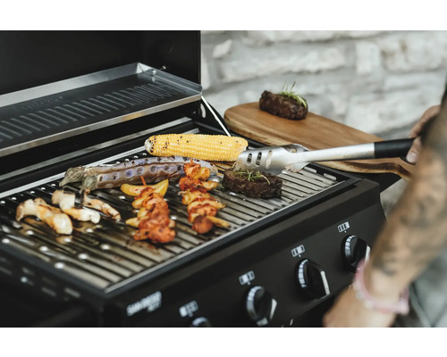 Grill mit Maiskolben, Fleisch, Grillspießen und Gemüse auf dem Grillrost, Grillzange zum Wenden des Grillguts