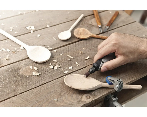 Mann schleift einen Holzlöffel mit einem Multifunktionswerkzeug auf einer Holzoberfläche. Verschiedene Holzlöffel und Schnitzwerkzeuge sind ebenfalls sichtbar.
