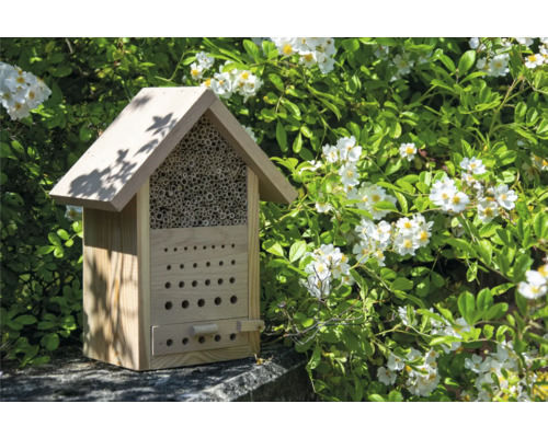Insektenhotel aus Holz mit verschiedenen Nistmöglichkeiten vor blühendem Strauch