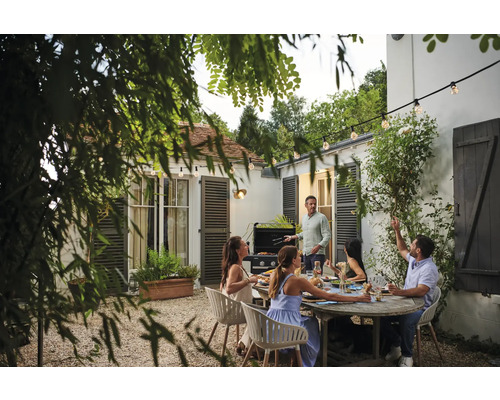 Gartenszene mit Grill, Tisch und sitzenden Personen unter einer Lichterkette