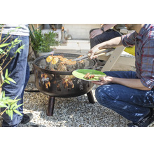 Feuerschale mit Grillrost im Garten, darauf Grillgut und eine Person mit Grillzange
