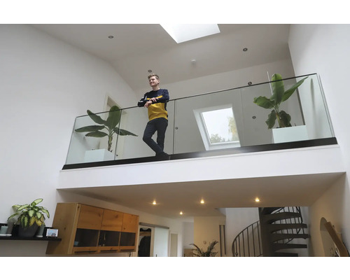 Ein junger Mann steht auf einer Galerie mit Glasgeländer in einem hellen, modernen Wohnraum mit Zimmerpflanzen und Oberlichtern.