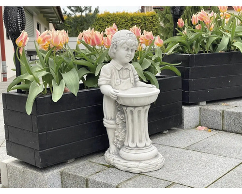 Dekorative Gartenfigur eines Jungen mit Vogeltränke, platziert in einem Außenbereich mit Blumenkästen.