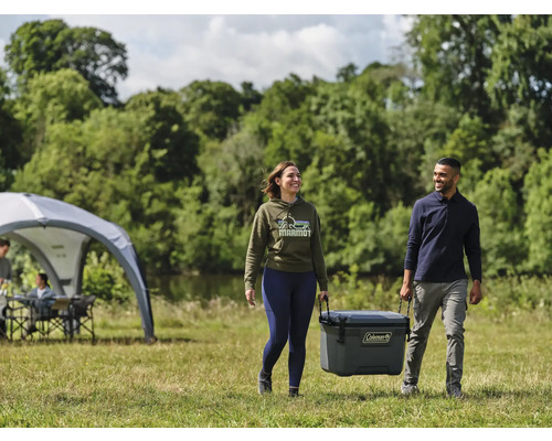 Ein Mann und eine Frau tragen eine Coleman Kühlbox in einem Park mit einem Zelt und Bäumen im Hintergrund.