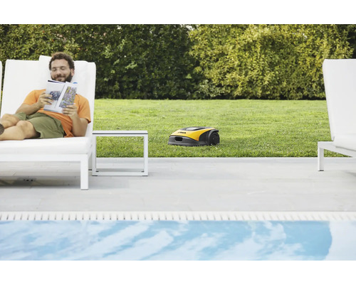 Ein Mann liest ein Buch auf einer Sonnenliege, während ein Rasenmähroboter den Rasen mäht.