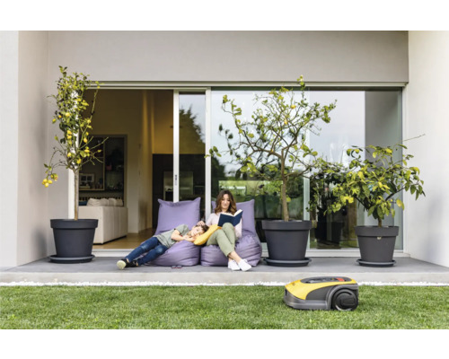 Eine Frau liest ein Buch, während ein Kind auf einem Sitzsack liegt, mit Zitronenbäumen in Töpfen und einem Rasenmähroboter auf dem Rasen.