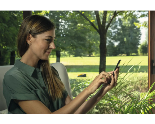 Eine Frau bedient ein Mobiltelefon mit einem Rasenmähroboter im Hintergrund.