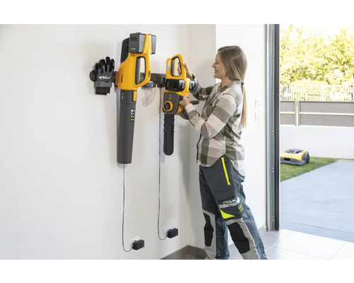Szene mit einer Frau, die eine Kettensäge an einer Wandhalterung befestigt, zusammen mit einem Laubbläser, Handschuhen und einem Rasenmähroboter im Hintergrund