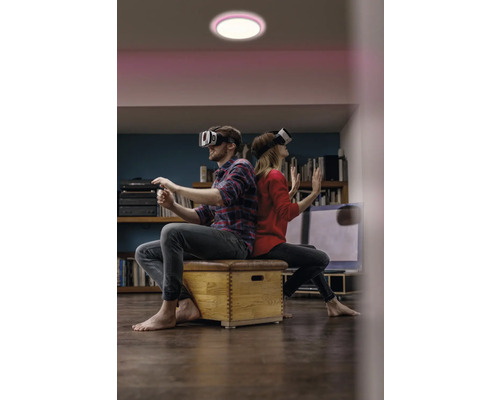 Mann und Frau sitzen mit Virtual-Reality-Brillen Rücken an Rücken auf einer Holzkiste unter einer Deckenleuchte.