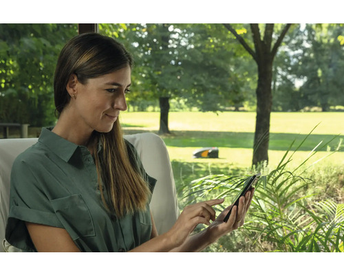 Frau bedient Rasenmähroboter per Smartphone im Garten.