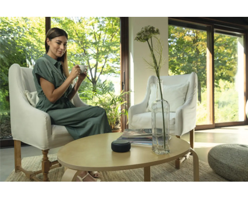 Frau mit Tasse sitzt in einem Sessel. Auf dem Tisch steht ein Lautsprecher