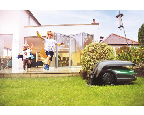 Zwei Kinder mit Superheldenkostümen rennen auf einen Rasenmähroboter im Garten zu.