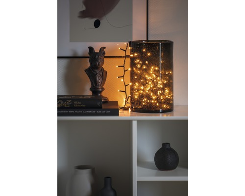 Dekoratives Arrangement mit Lichterkette in Glasvase, Skulptur und Büchern auf einem Regal