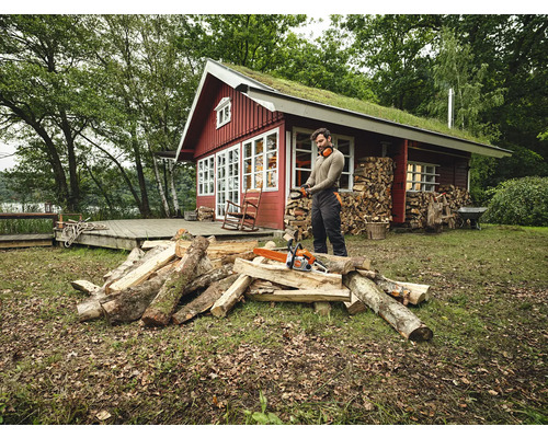 Mann mit Kettensäge beim Holzspalten vor einem Holzhaus mit Gründach.