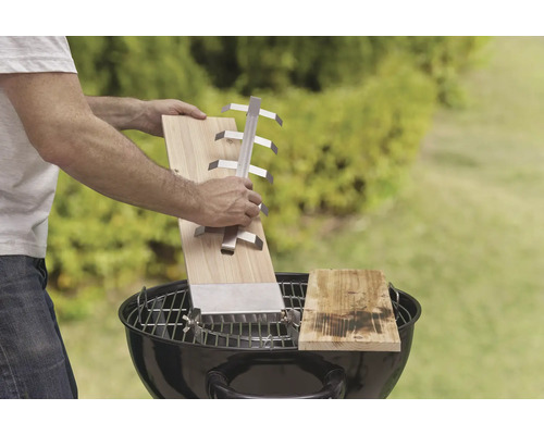 Szene mit Mann, der ein Grillbrett mit Halter über einem Holzkohlegrill hält, im Freien.