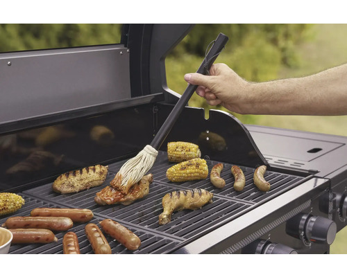 Mann bestreicht Fleisch mit einer Marinade auf einem Grill mit Maiskolben und Würstchen.