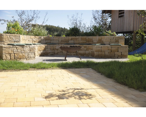 Gartenszene mit Steinmauer, Sitzgelegenheit mit Kissen und Feuerschale auf gepflasterter Fläche