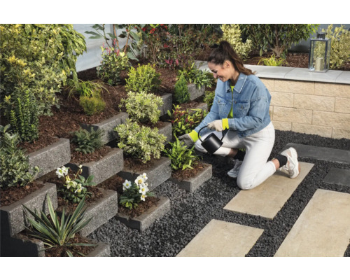 Gestalteter Garten mit gestuften Pflanzgefäßen, bepflanzt mit verschiedenen Pflanzen, und einer Frau, die die Pflanzen mit einer Gießkanne gießt.