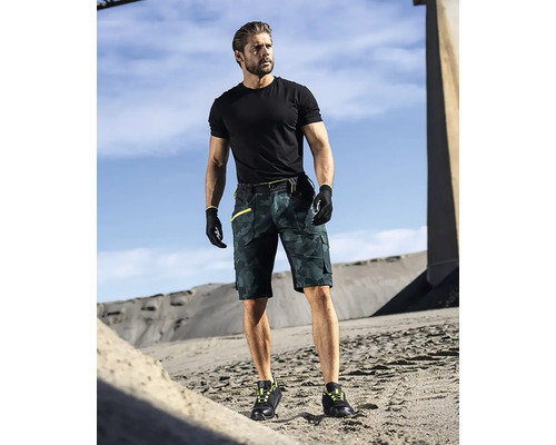 Ein Mann trägt ein schwarzes T-Shirt, Arbeitshandschuhe, eine kurze Arbeitshose mit Tarnmuster und Arbeitsschuhe.