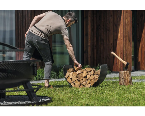 Mann befüllt Brennholzregal mit Holzscheiten neben einem Hackklotz mit Axt im Garten.