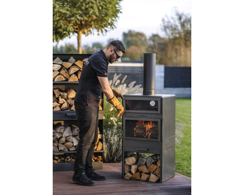Mann mit Schutzhandschuhen bereitet einen Cook King Grillofen im Garten vor. Holzstapel neben dem Ofen.
