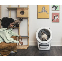 Eine Frau hockt neben einem Katzenkletterbaum und einem automatischen Katzenklo in einem Raum, in dem Katzenprodukte ausgestellt sind.
