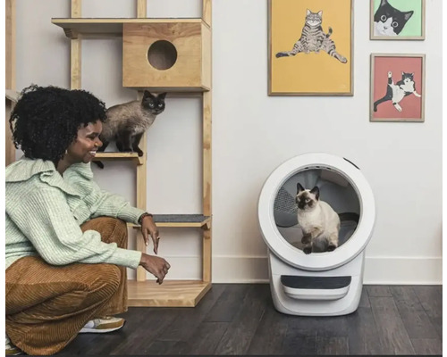 Eine Frau hockt neben einem Katzenkletterbaum und einem automatischen Katzenklo in einem Raum, in dem Katzenprodukte ausgestellt sind.