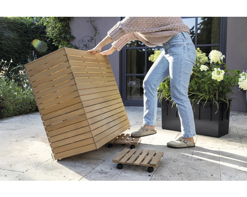 Eine Person bewegt einen Pflanzkübel aus Holz auf einem Rollbrett im Garten.
