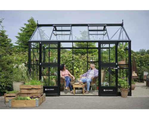 Gewächshaus JULIANA Jubii 60 10,9 m² Sicherheitsglas 3 mm 296 x 368 cm schwarz Gewächshaus mit zwei Personen beim Entspannen im Inneren, umgeben von Pflanzen und Gemüsebeeten.