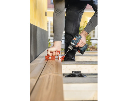 Eine Person montiert eine Holzterrasse mit einem Abstandhalter, einem Akkubohrer und Terrassenschrauben.