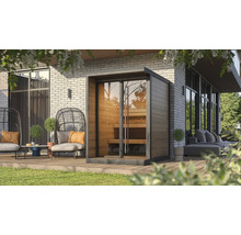 Moderne Außensauna aus Holz mit Glasfront und Innenbänken auf einer Holzterrasse mit Gartenmöbeln vor einer Ziegelwand.