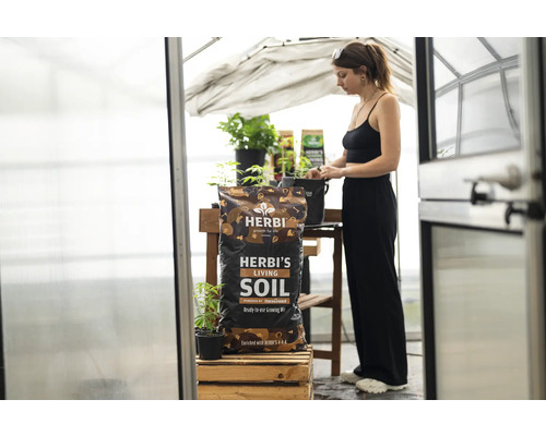 Ein Sack Herbi's Living Soil in einem Gewächshaus mit einer Frau im Hintergrund.