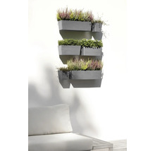 Mehrere graue Wand-Blumenkästen an einer hellen Wand, bepflanzt mit Heidekraut und Grünpflanzen über einem Gartensofa in einem hellen Außenbereich.