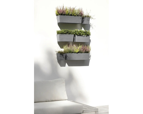 Mehrere graue Wand-Blumenkästen an einer hellen Wand, bepflanzt mit Heidekraut und Grünpflanzen über einem Gartensofa in einem hellen Außenbereich.