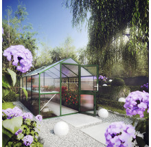 Gewächshaus mit grünem Rahmen und transparenten Hohlkammerplatten in einem hellen Garten mit Pflanzen, Kies und lila Blumen.