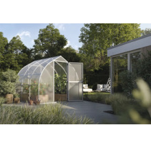 Hell gestalteter Garten mit modernem, gewölbtem Gewächshaus aus Metall und Polycarbonat. Im Inneren wachsen Gemüsepflanzen auf einer Holzterrasse.