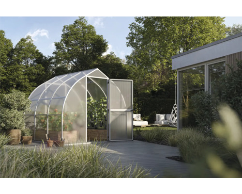 Hell gestalteter Garten mit modernem, gewölbtem Gewächshaus aus Metall und Polycarbonat. Im Inneren wachsen Gemüsepflanzen auf einer Holzterrasse.