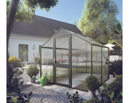 Freistehendes Gewächshaus mit anthrazitfarbenem Aluminiumrahmen und Hohlkammerplatten in einem hellen Garten neben einem Wohnhaus.